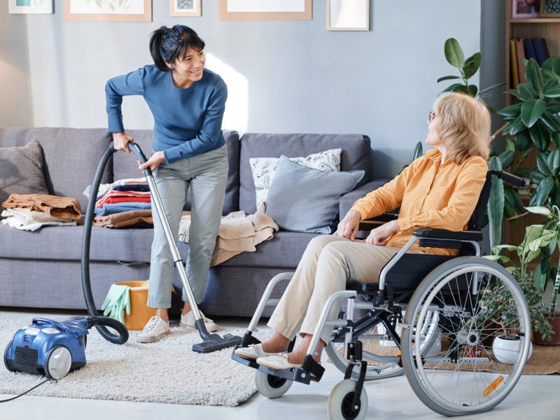 Housemaid using vacuum cleaner to clean the carpet in the room, she helping senior woman who using wheelchair