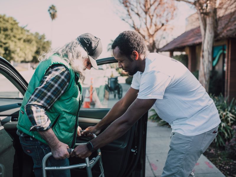 Elderly man holding a walker while being aided out of a car by another man